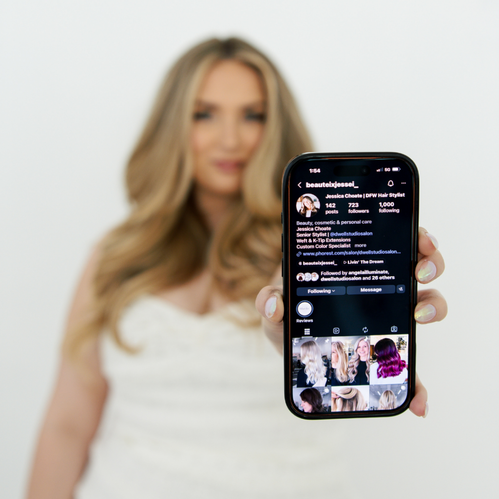 Woman holding a smartphone displaying an Instagram hair styling profile at Dwell Studio Salon in Fort Worth salon.